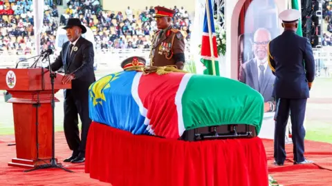 Reuters A coffin draped in the Namibian flag stands on a stage as a man in a black suit speaks at a lectern.