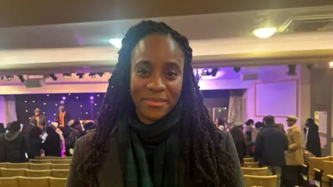 Obehi Orukpe is seen looking direct at the camera in the church. Vigil attendees are seen standing in the background as well as empty seating.