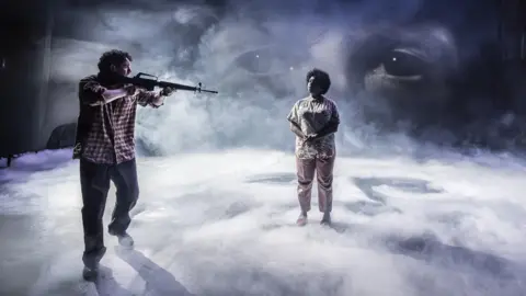 Marc Brenner Robbie O’Neil (Brendan) pointing a gun at Anita Reynolds (Caroline) amid swirling dry ice and in front of a giant pair of eyes in The Assassination of Margaret Thatcher at Liverpool Everyman