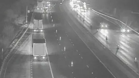Cones can be seen on a black and white image of the motorway closing off some lanes. Traffic is driving on the remaining lanes. 