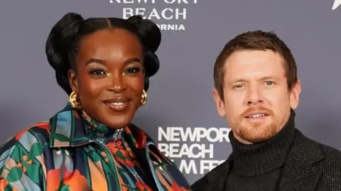 PA Media Wunmi Mosaku and Jack O'Connell as they pose together at a presscall. She wears a bright mulit-coloured top and jacket, while he wears a black poloneck jumper and jacket.