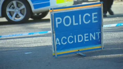 BBC A police accident sign, blue with white lettering, on a road in front of white and blue police tape. The steering wheel of a police car is visible in the background, as are the feet of an officer.