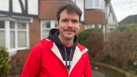 Josh McLaughlin/BBC James Roberts, who has brown hair and a moustache. and is wearing a red raincoat and dark hoodie. He is standing outside a house in Hove.