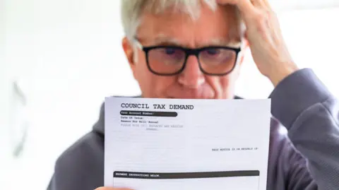 Portrait of a senior man in his 70s checking his council tax bill at home in his office. He has a worried expression and touches his face with his hand while looking at the bill
