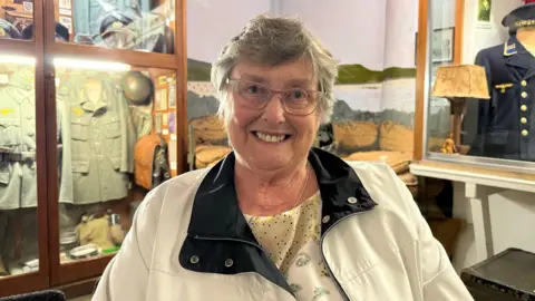 A headshot of a woman with short grey hair, glasses smiling at the camera. She's sat in from of cabinets containing war artefacts and wearing a cream jacket with a navy collar.