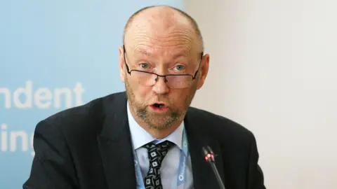 PA Media A man with a brown beard and glasses. He is wearing a black jacket and a blue shirt with a black tie
