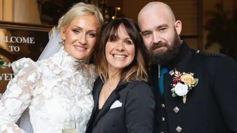 Hannah Howie Hannah in a white wedding dress posing alongside Carol Smillie - who has dark hair and is wearing a black jacket - and Hannah's husband, who is bald and wearing a kilt suit jacket. All three are smiling. 