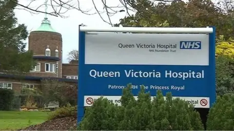 A blue and white sign reading The Queen Victoria Hospital NHS Foundation Trust. It is in front of a brown building surrounded by grass and trees.