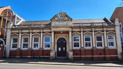 Rushden Library, an old stone built building with a door in the middle and four windows either side. A road is in front of it and a glass structure to the left. It is an ornate building. 
