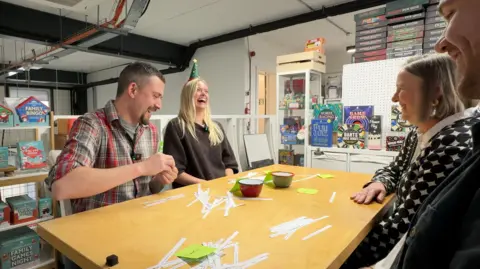 Two men and two women test a number of jokes on one another as part of Christmas cracker gag selection for 2026. The group sits around a table, three are smiling and one, a woman, is laughing heartily