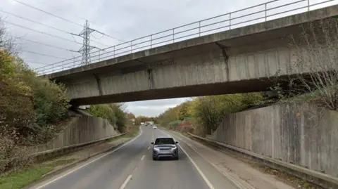 Google A concrete railway bridge over a modern bypass. Concrete walls support the bridge, but elsewhere, semi-mature trees line both sides of the route.