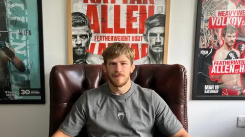 Cleah Hetherington/BBC A picture of Arnold Allen. He is sitting in a brown leather chair facing the camera. He has blond hair and a blond beard. He is wearing a grey t-shirt. Posters from his fights are placed on the wall behind him.