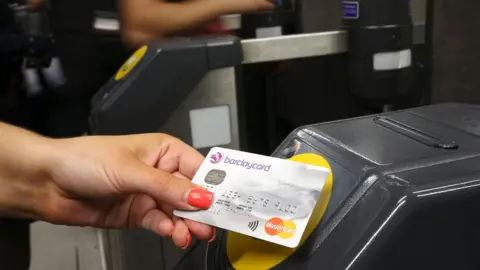 Commuter holds credit card to yellow Tube gate card reader