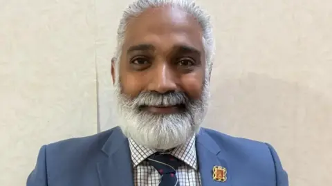 Lancashire County Council Dr Sakthi Karunanithi wearing a blue suit with a checked shirt with a large white beard. He stands in front of a wall with cream wallpaper. He is smiling.