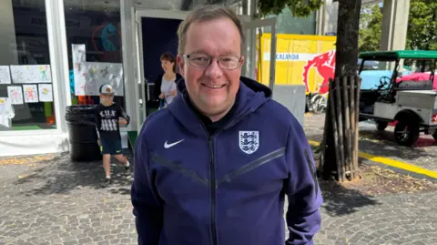 England fan Phillip Spakouskas. He is wearing a navy blue zipped jumper with the Coat of Arms of England on it, and has short brown hair and glasses. He is standing on a cobbled street with a tree and family behind him.