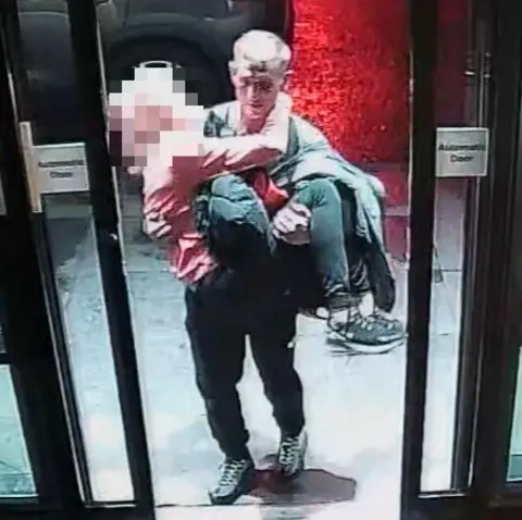 COPFS CCTV footage of Michael Harvey walking through hotel sliding doors carrying Carol O'Reilly in his arms. Her face has been blurred