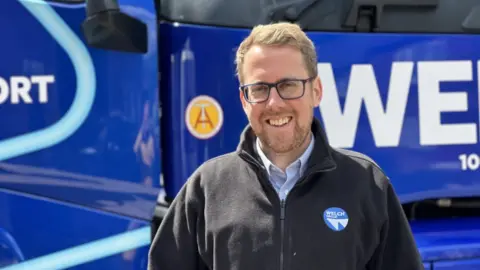 A man with short blonde hair and a beard, wearing dark rimmed glasses and a dark zip-up jacked in front of a bright blue vehicle