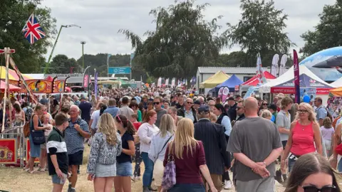 Crowds of people at Driffield Showground. There are stalls down either side and in the middle, hundreds of people are walking around and looking at the attractions.