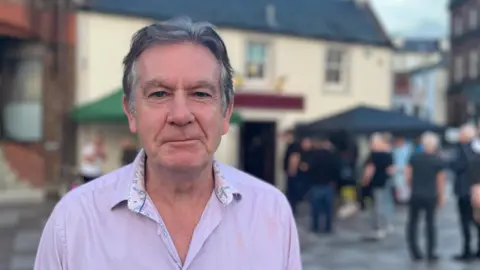 Stewart Solley, who has grey hair, is wearing an open-neck pink shirt and is standing in front of a popular live music venue in Dumfries