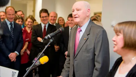 The Sir Bobby Robson Foundation Sir Bobby speaking into a microphone and looking to the left. He is wearing a grey suit with a pink tie and blue shirt. To the right of him is a woman with short brown hair. To the left is a crowd of people which includes Alan Shearer and Fabio Capello who are both wearing suits and smiling. Hospital staff are behind them in red uniforms. 