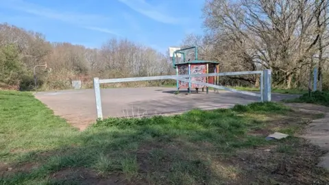 A park. There is a tarmac square in the middle which has a shelter on it that is covered in graffiti. Attached to the shelter is a sports hoop. Around the tarmac is patchy green grass with areas of mud. There is a gate on one side of the tarmac square.