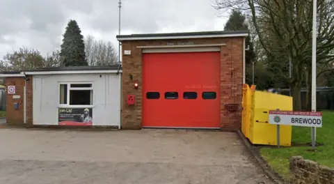 A small fire station with a red garage door. A large driveway is outside with fire service sign saying 'Brewood'. Behind it are two yellow boxes and some grass. A white door is also visible with fire service poster which says 'Join Us'. 