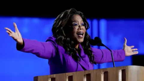 Oprah Winfrey, wearing a purple suit jacket, raises her arms as she gives a speech at the Democratic National Convention in Chicago