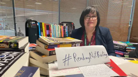 BBC Denise Jones from Liverpool library service surrounded by books. She has short dark hair and is wearing glasses. A sign in front of her reads #ReadingnotRioting
