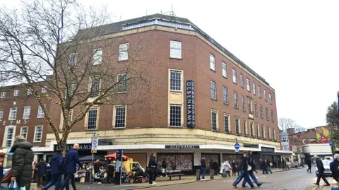 A picture of the Debenhams building on a busy shopping day. The building is 5 or 6 storeys tall. A number of people are walking past it or crossing the road in front of it.