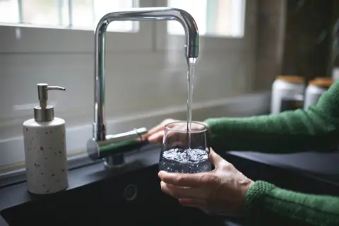 Getty Images Hand pouring a glass of water from a tap, over a sink, soap dispenser pictured beside it, can see persons arms in green wool jumper