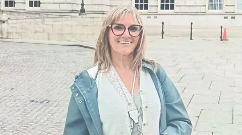 A middle aged woman with long grey/blond hair and glasses stands in the grounds of Belfast City Hall