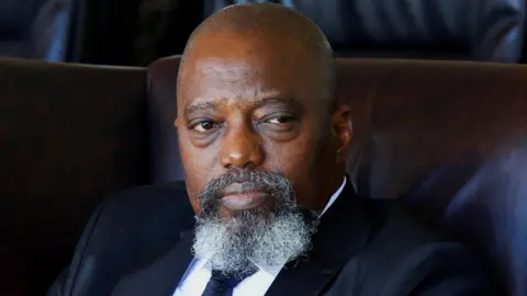 Joseph Kabila, with a grey goatee beard, in a black suit and tie sitting in a leather chair.