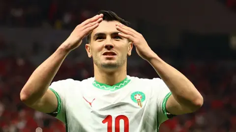 Brahim Diaz with hands raised to head height wears the Morocco green and white shirt with number 10 on the front
