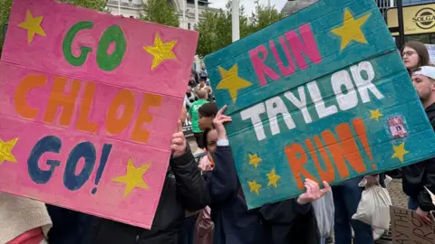 Great Birmingham Run Spectators hold placards saying Go Chloe Go! and Run Taylor Run! They are painted in bright colours and decorated with stars. The people with the banners are holding them in front of their faces. Crowds are visible behind them.