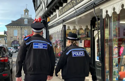 A police community support officer and another person in a black uniform with the word police on their back are on the pavement. They have their backs to the camera and buildings are behind them on the image.