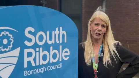 Councillor Clare Hunter with long blond hair wearing a black blazer stands at the side of a blue and white sign for 'South Ribble Borough Council'.