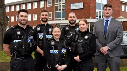 Two women and three men in black police uniform, and a man in a grey suit, standing and smiling in front of a brick building with Thames Valley Police written above the door.
