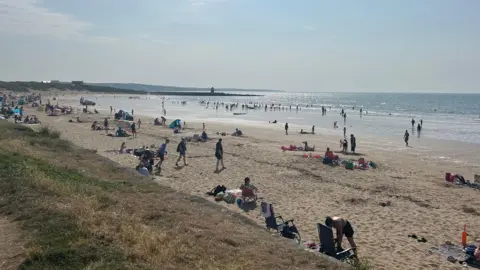 BBC Beach with people on the sand and sea shore as well as swimming in the sea. The sun is shining and everyone is in swimwear or summer clothing. Some people have surf boards.