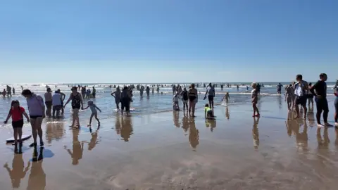 A view of a beach, with swimmers and people with body boards standing on the sand. In the distance you can see people in the water