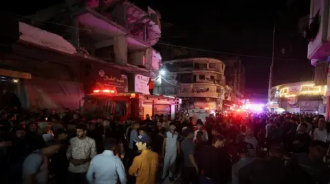 Large crowds gathered at the scene of the attack, with emergency vehicles flashing beside damaged buildings