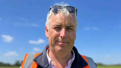 Simon Hutton has grey hair and sunglasses, he's also wearing an orange high vis jacket. The bright blue sky with a few dotted clouds is behind him.