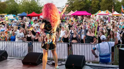 Pride in Gloucestershire A performer dressed in colourful clothes stands with their back to the camera on stage, looking out to a sea of fans below.