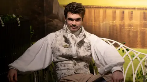 Jane Austen Centre A man dressed as Mr Darcy in 18th Century clothes, reclining on an iron garden bench with a painted background of a lake behind him  