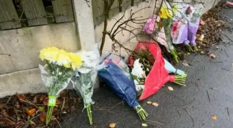 7 bunches of flowers in plastic wrappings left propped on a wet pavement against a white stone wall next to a road.