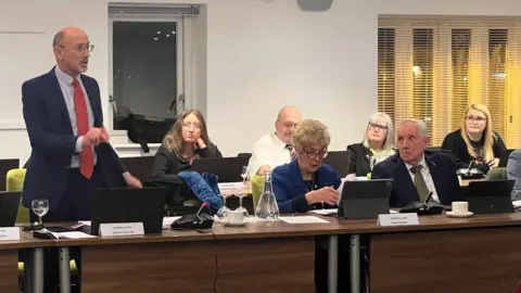 Carmelo Garcia Labour councillor Terry Pullen speaking a council meeting, standing behind a long wooden desk beside other seated council members. They are in a white room at night, with name cards in front of them. Pullen is wearing glasses, a dark suit and red tie, gesturing with his hands.