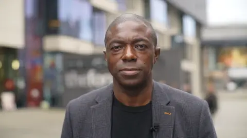 Jamie Niblock/BBC Bayo Alaba is in a black top and a faintly-striped dark-coloured suit jacket over the top. He is looking at the camera, standing outside.