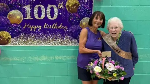 Gianni Epifani There is a purple wall hanging sign reading "100th happy birthday" with balloons and sparkles. To the right is Elizabeth Corran with her arm around Lillian Pike. Elizabeth is handing her a basket of flowers and Lillian is wearing a gold sash.