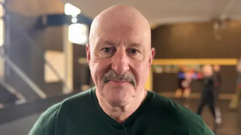 A close-up image of Martin O'Neill. He is an older, bald man with a grey moustache. He is smiling at the camera with his mouth closed. Behind him are other participants of the Irish stick fighting class, but the background is blurred.