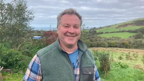 Matt, a middle aged man with curly grey hair, wearing a blue check shirt and a fluffy green gillet. He is stood in a field with more grassy fields and hills behind him and is smiling as he looks at the camera.