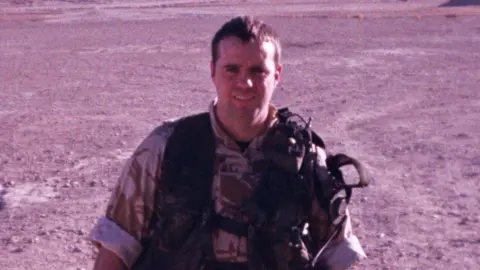 Kevin Russell looks directly at the camera standing in a desert. He is wearing Army combat gear. He has brown hair. 